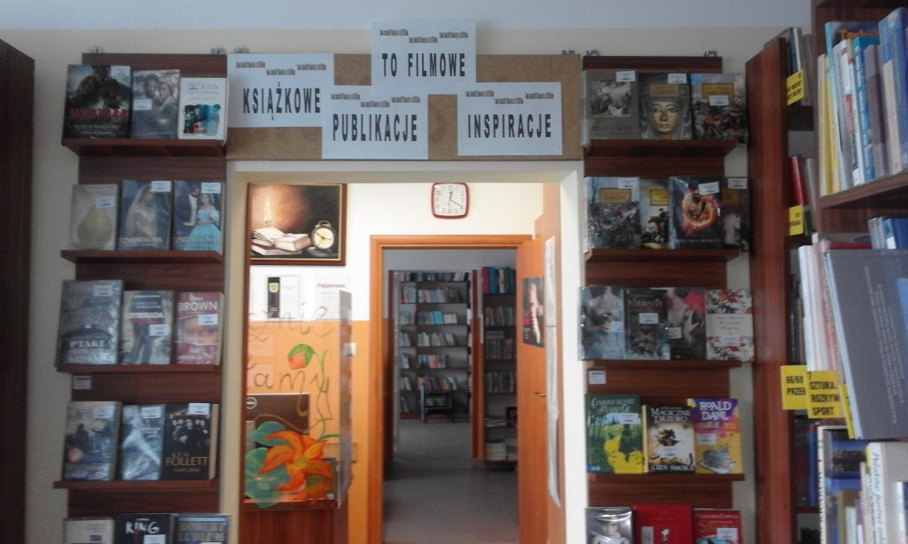 Wnętrze biblioteki. Po lewej i prawej stronie od wejścia wystawka filmowych ksiażek. W dalszej perspetywie od lewej stronie widać stanowisko bibliotekarz w dalszym pomieszczeniu półki z książkami.