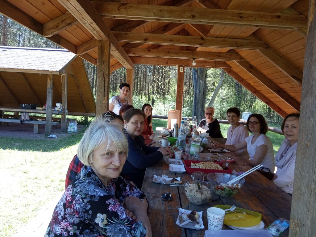 Przy ławie w lesie siedzą kobiety i jedzą posiłek.